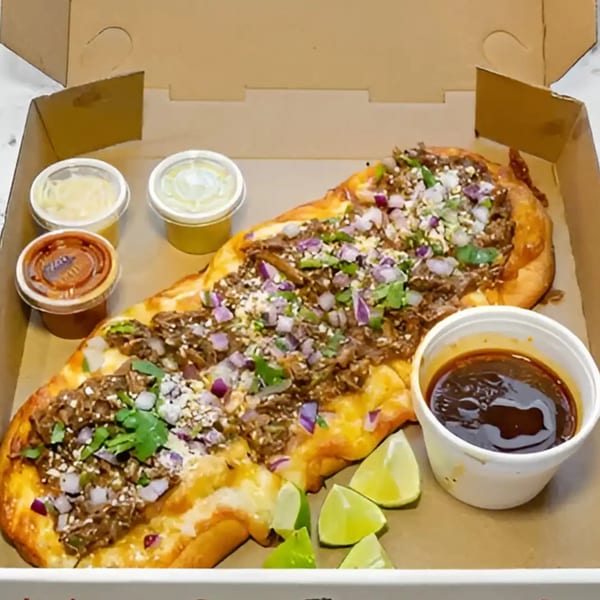A large Mexican pizza with meat, onions, cilantro, and cheese, served with lime wedges and sauces in a box.