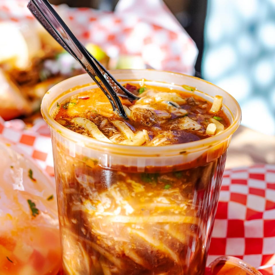 Birria consommé with noodles and meat in a clear cup with a red checkered liner.